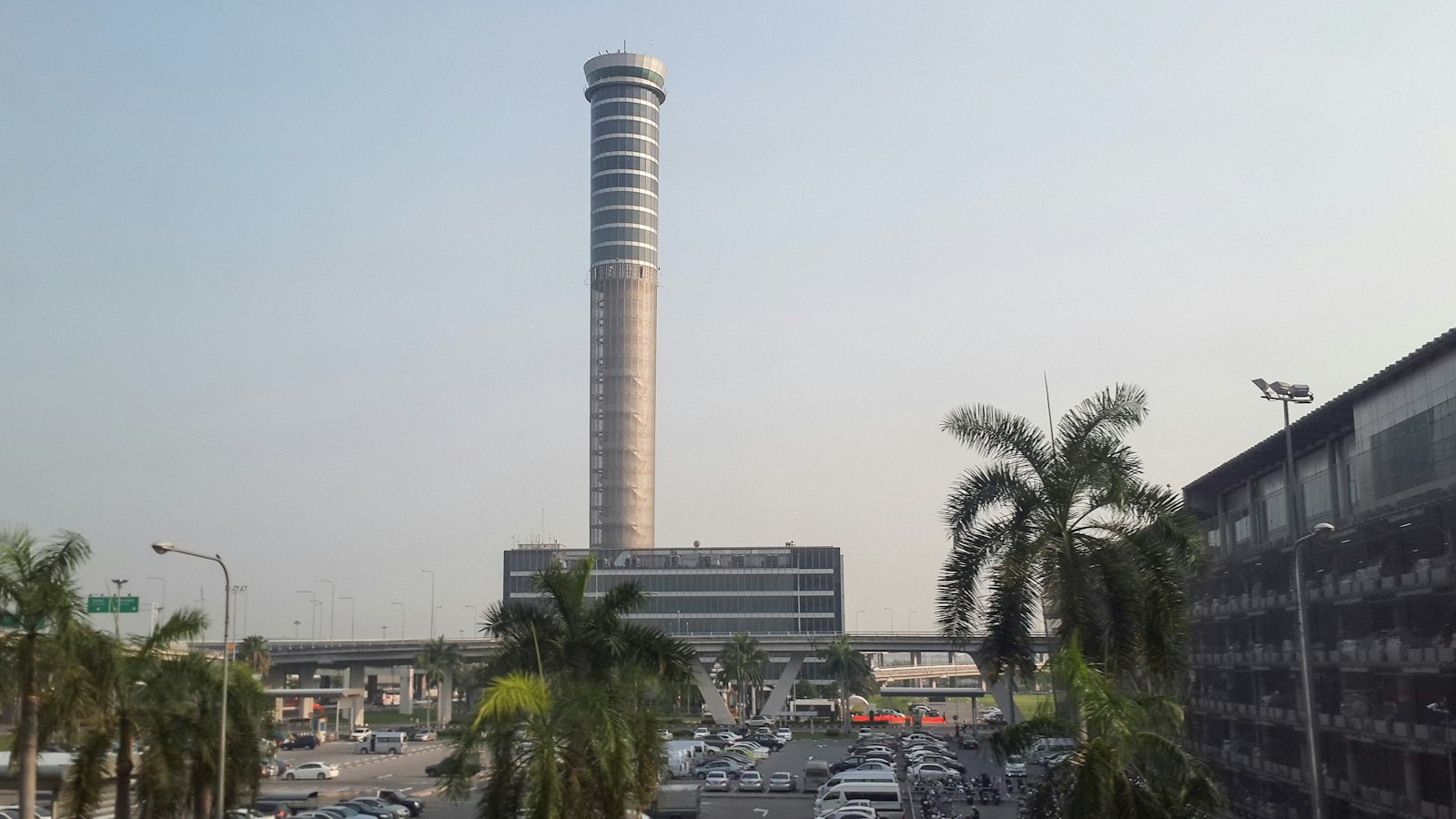 Air traffic control tower at Bangkok Suvarnabhumi Airport rising above palm trees and the airport car park